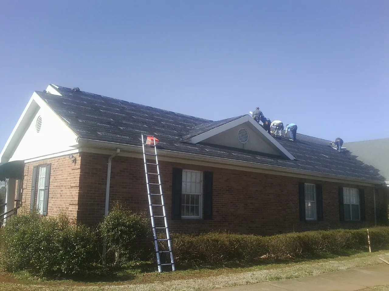 Crew tearing shingles off a brick ranch roof with a ladder set — Metal Roof Repair work by our crew in Bentleyville