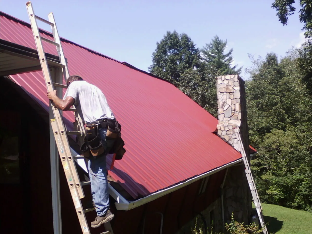 Roofer on a ladder beside a red metal roof and stone chimney — Soffit & Fascia Repair by Roofing Brothers in Bentleyville